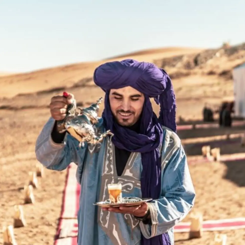 A man in a purple turban pouring tea from a height at a desert camp